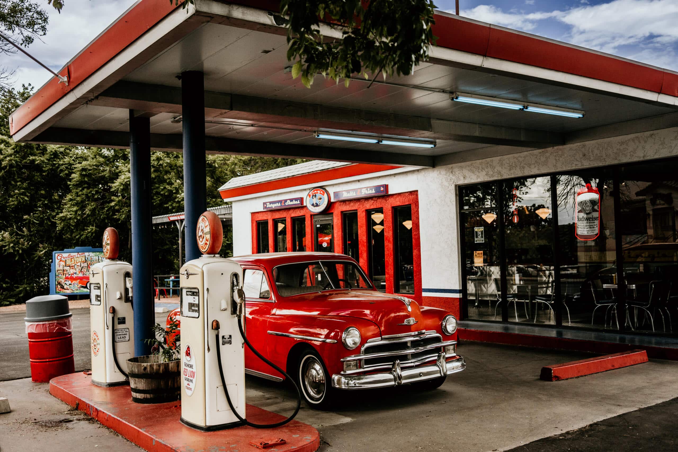 Vintage red car parked at retro 1950s-style gas station with old pumps and storefront.