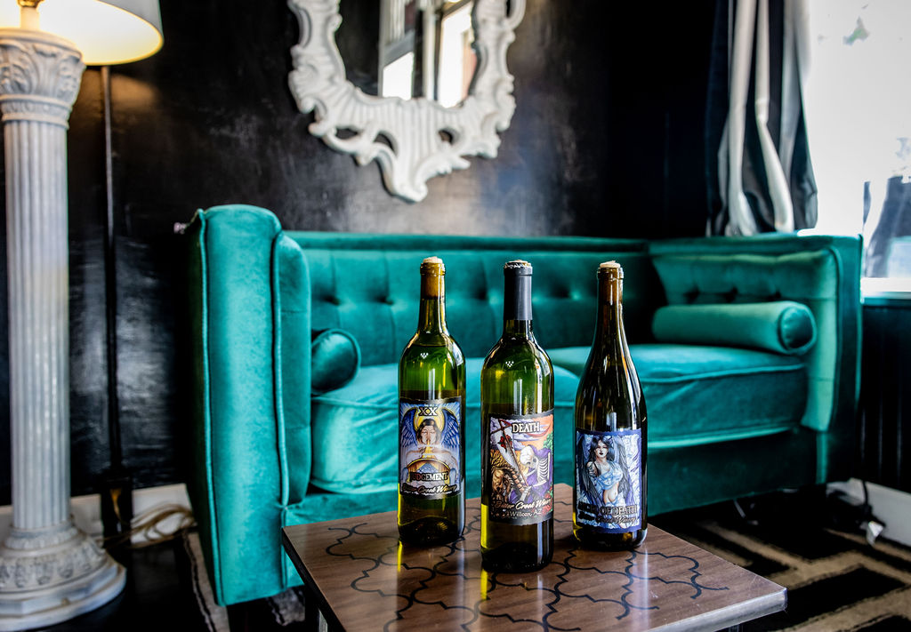 Three bottles of wine labeled 'Death' on a table near turquoise velvet furniture.
