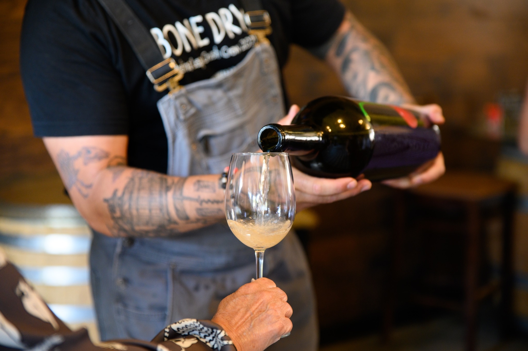 Bartender pouring white wine into glass for tasting, wearing apron and tattoos.