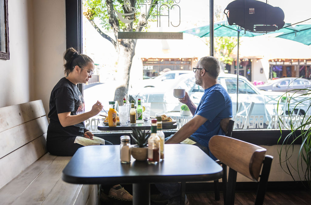 Two people sitting at café table enjoying drinks and food outdoors.