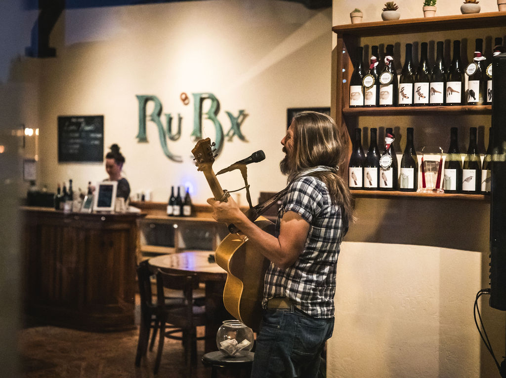 Man playing acoustic guitar at a cozy bar named RuBx with bottles on display.