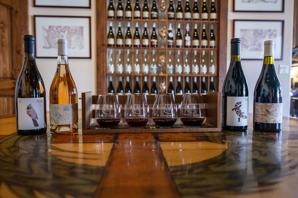 Wine tasting setup with four bottles and five glasses on wooden table.