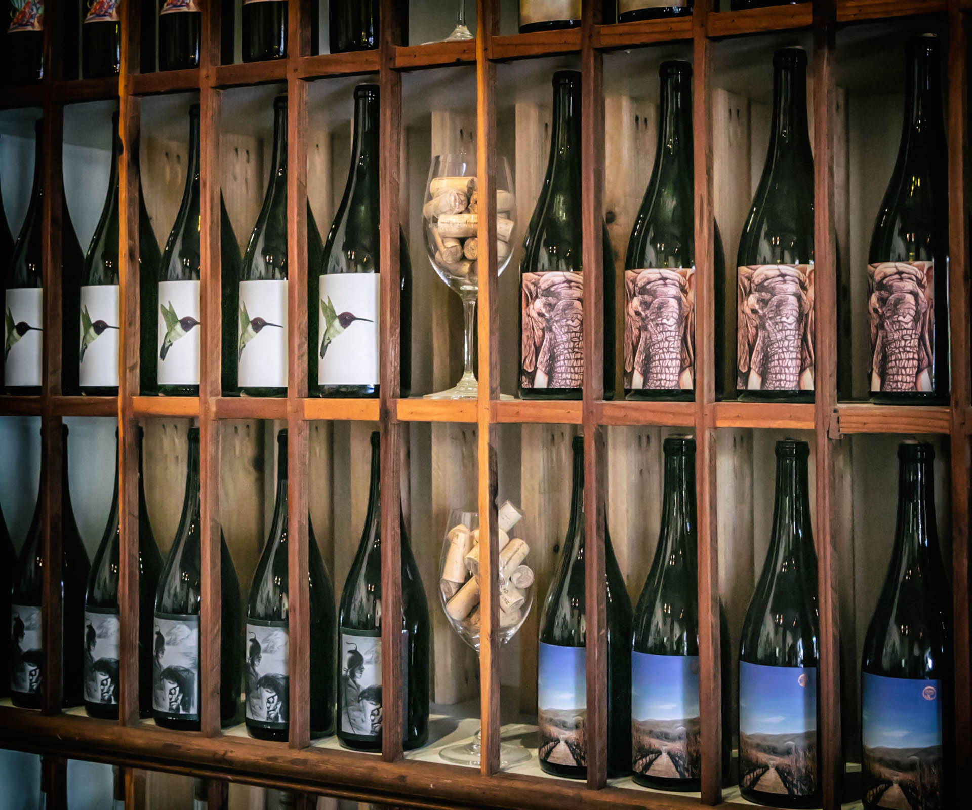 Wooden shelf displaying various labeled wine bottles and glassware.