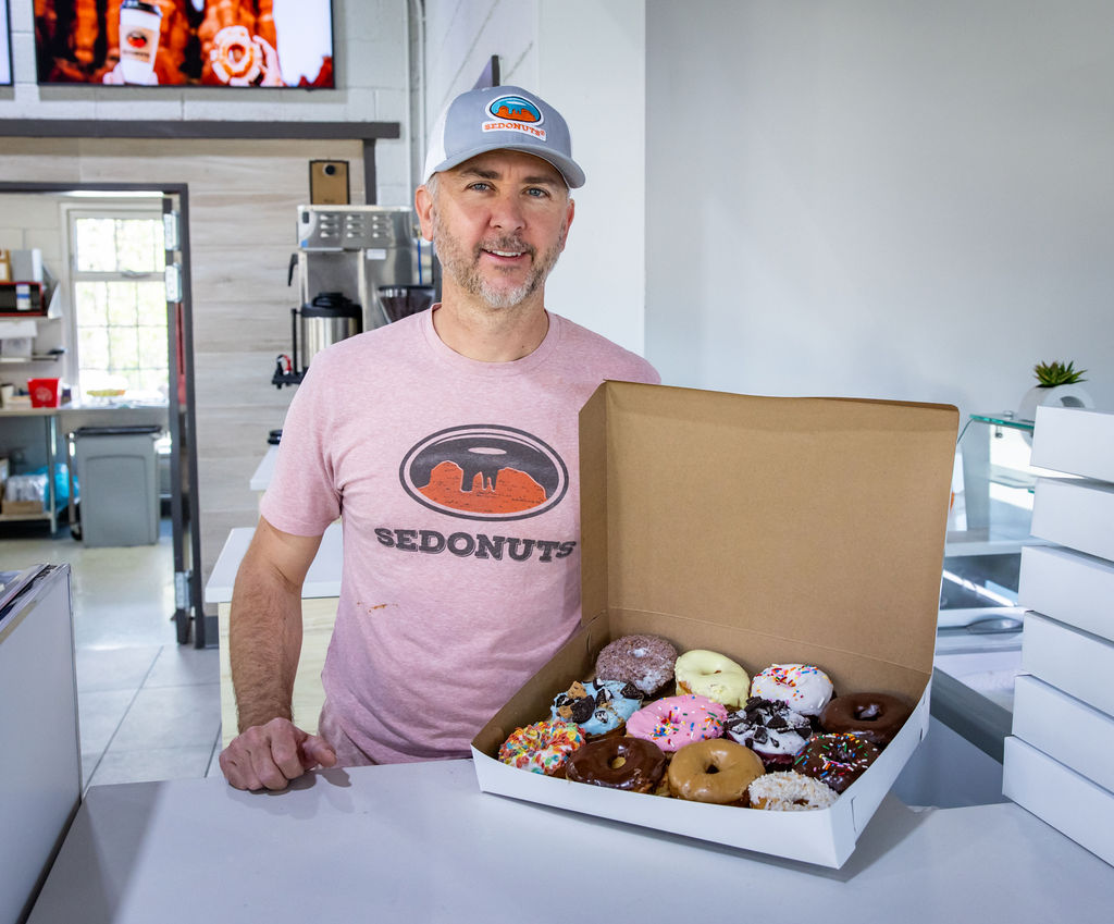 Man in Sedona Donuts cap and shirt holding box of assorted colorful donuts in kitchen.