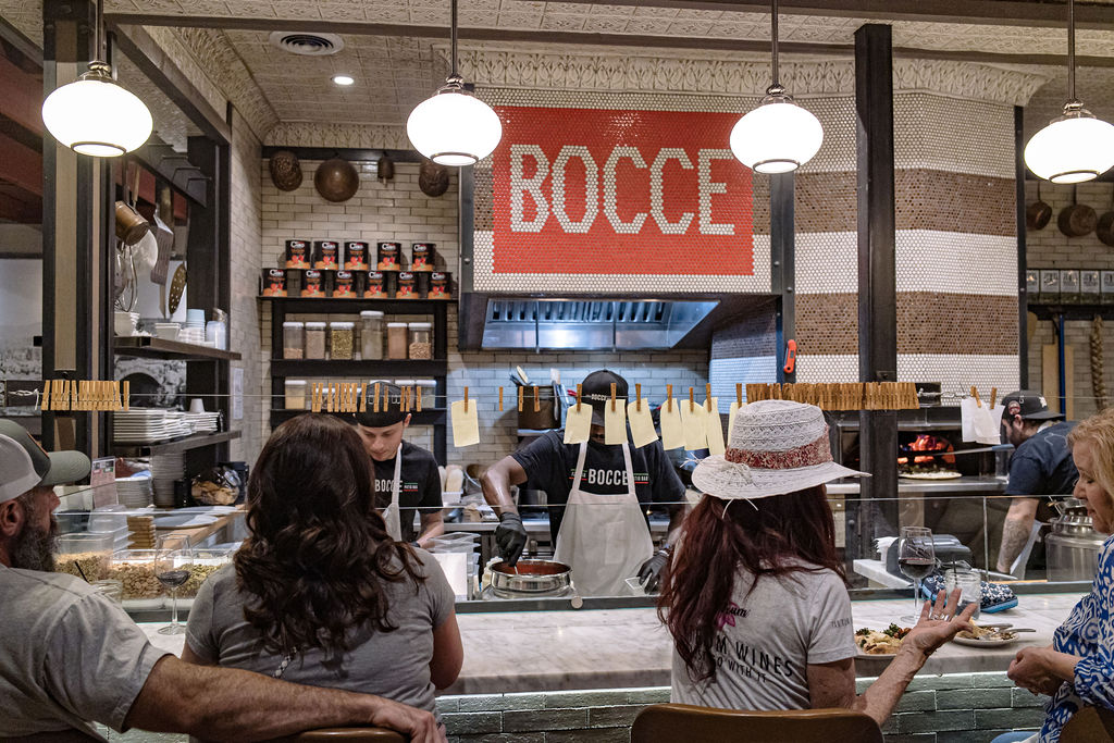 Customers dining at Bocce restaurant with chefs preparing food in a bustling kitchen.