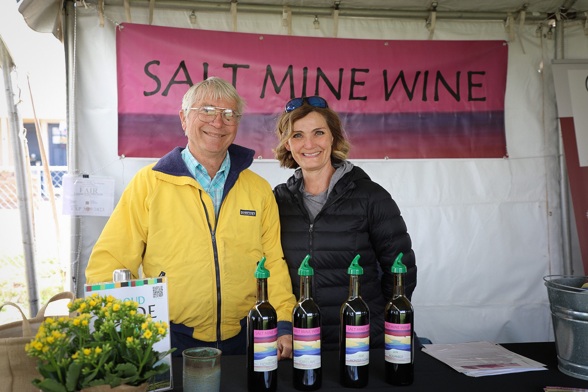 Two people smiling at Salt Mine Wine booth with wine bottles on display.