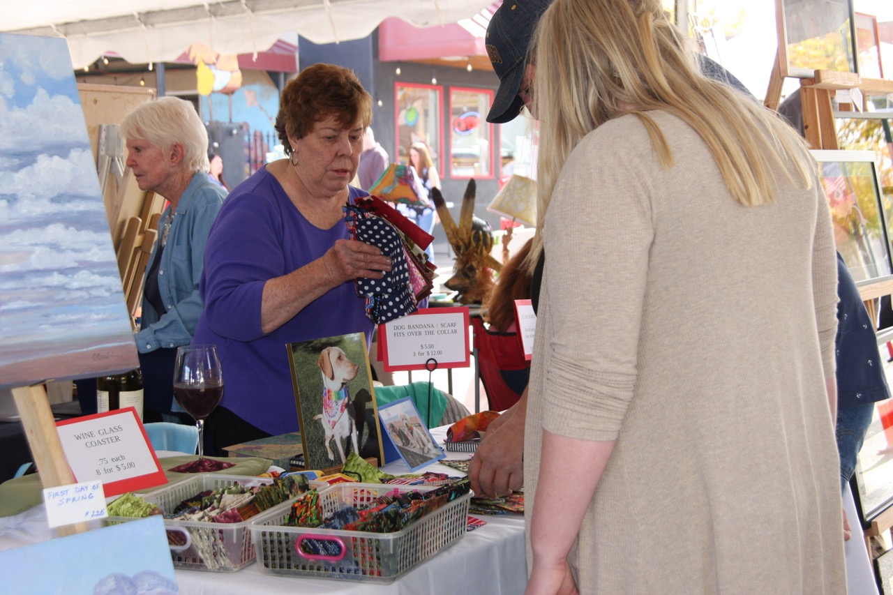 Women browsing pet bandanas, wine glass coasters at outdoor artisan market stall