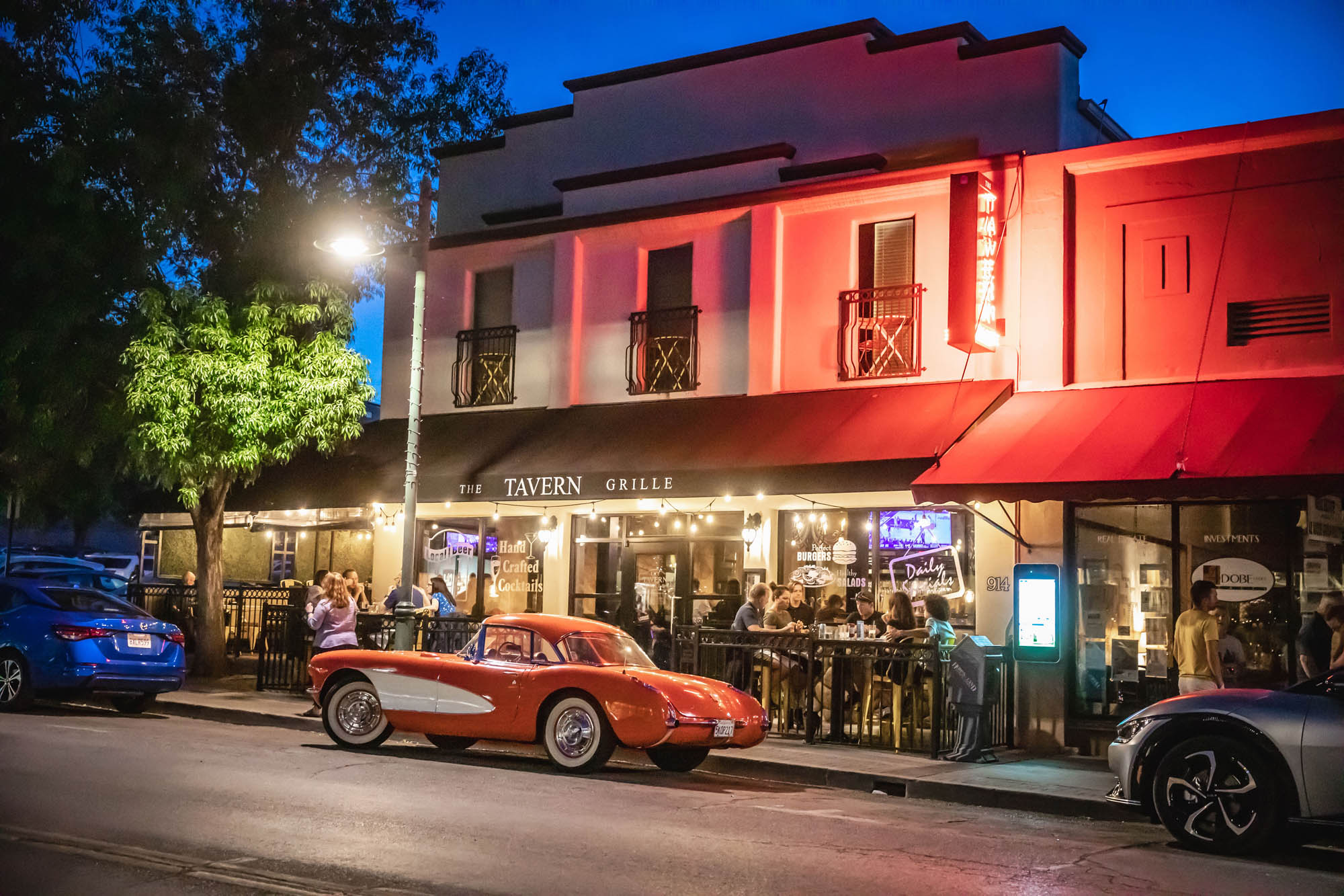 Vintage red car parked outside lively evening tavern with patrons dining