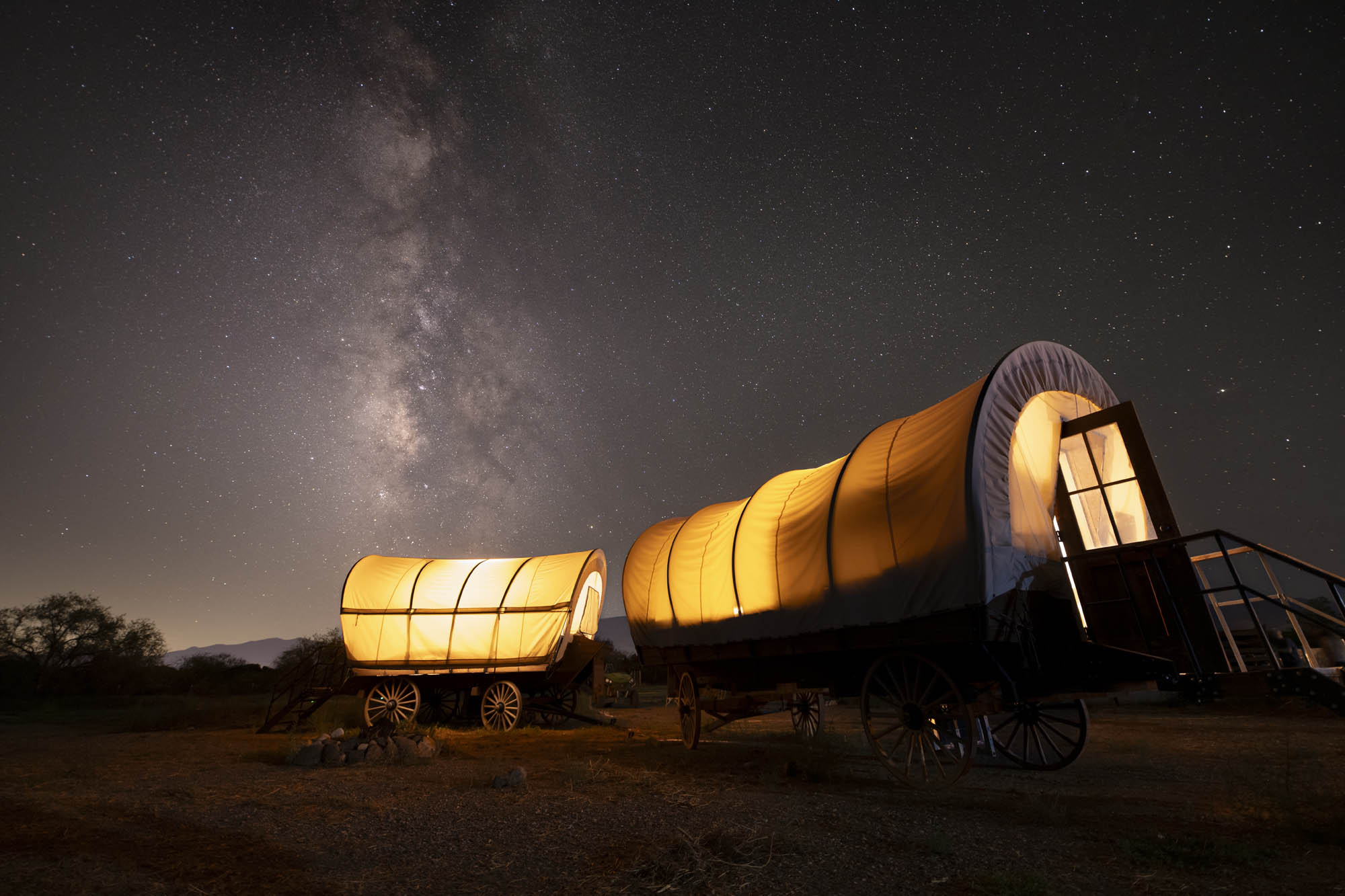 Historic wagon under Milky Way galaxy night sky, illuminated by warm indoor light.