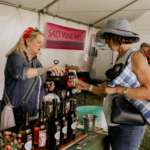 Two women sampling Salt Mine Wine at an outdoor market booth with various bottles displayed.