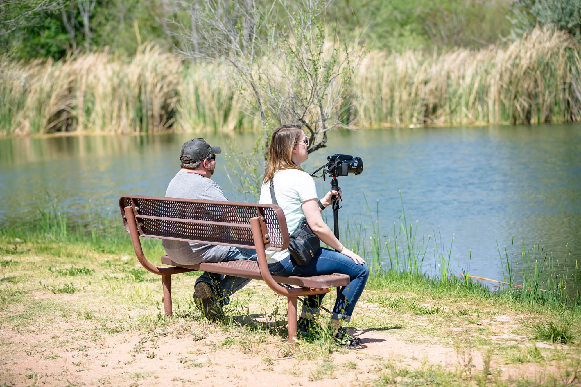Two people sitting on bench near water while filming outdoors